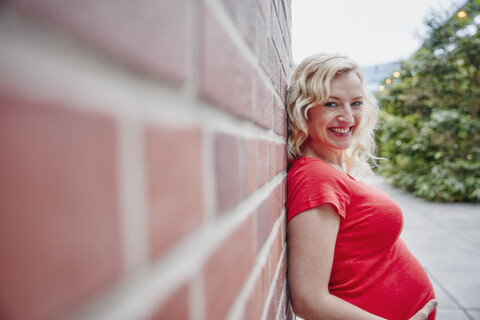 Porträt einer lächelnden schwangeren Frau, die sich im Freien an eine Backsteinmauer lehnt, lizenzfreies Stockfoto