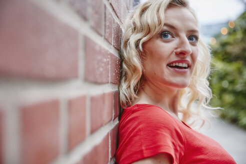 Porträt einer blonden Frau, die sich im Freien an eine Backsteinmauer lehnt - RHF02079