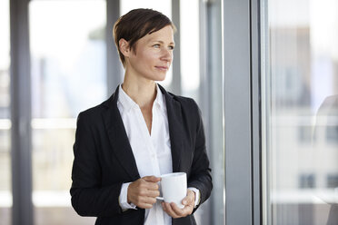 Businesswoman in office with cup of coffee looking out of window - RBF06357