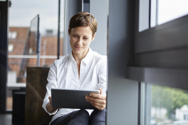 Geschäftsfrau sitzt im Büro am Fenster und benutzt ein Tablet - RBF06379