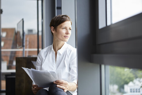 Geschäftsfrau sitzt im Büro mit Dokumenten und schaut aus dem Fenster - RBF06380