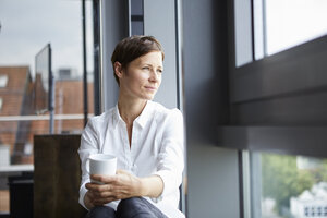 Geschäftsfrau sitzt im Büro mit einer Tasse Kaffee und schaut aus dem Fenster - RBF06381