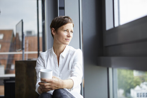 Businesswoman sitting in office with cup of coffee looking out of window - RBF06381