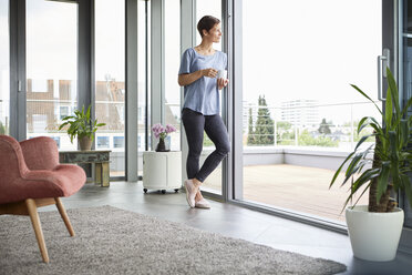 Woman at home with cup of coffee looking out of balcony door - RBF06385