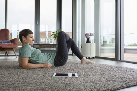 Woman lying on the floor at home with tablet looking out of balcony door stock photo