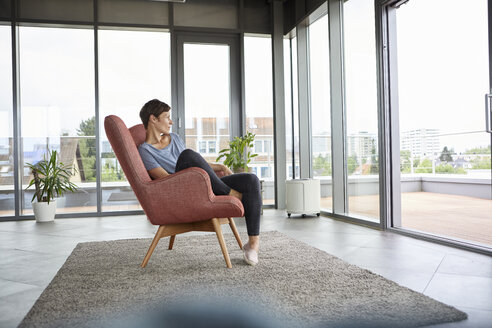 Woman sitting in armchair at home looking out of balcony door - RBF06393