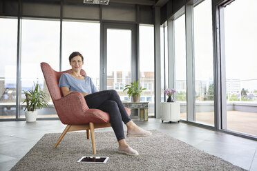 Porträt einer Frau, die zu Hause in einem Sessel sitzt und ein Tablet neben sich hat - RBF06394