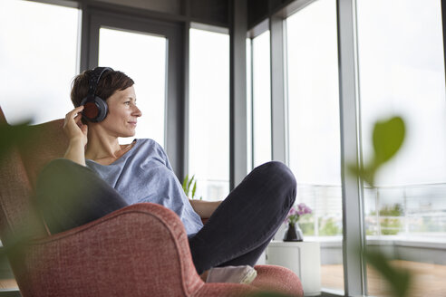 Frau sitzt zu Hause im Sessel und hört mit Kopfhörern Musik - RBF06395
