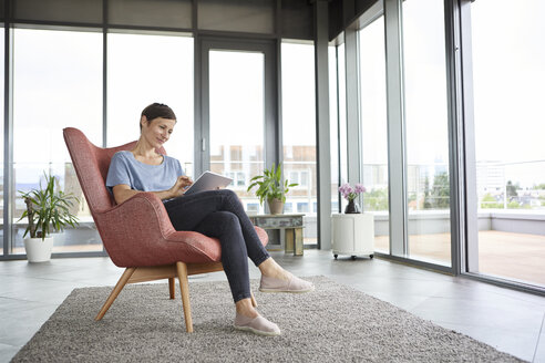 Frau sitzt in einem Sessel zu Hause und benutzt ein Tablet - RBF06399