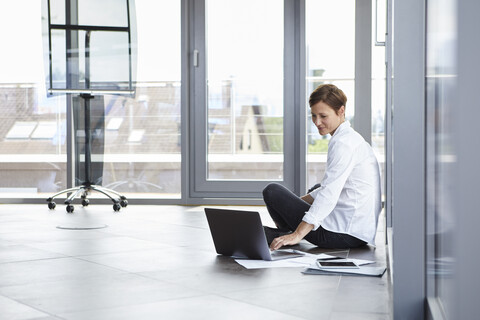 Geschäftsfrau sitzt auf dem Boden im Büro und benutzt einen Laptop, lizenzfreies Stockfoto