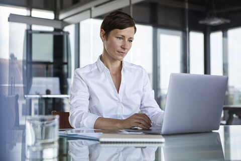 Geschäftsfrau sitzt am Glastisch im Büro und benutzt einen Laptop, lizenzfreies Stockfoto