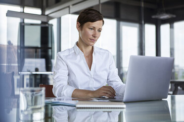 Businesswoman sitting at glass table in office using laptop - RBF06407