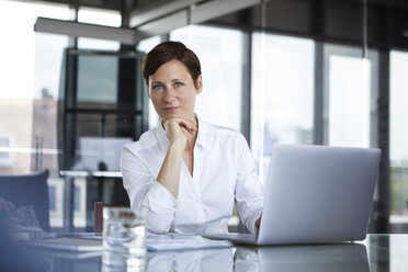 Portrait of confident businesswoman sitting at glass table in office with laptop - RBF06408