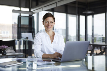 Porträt einer lächelnden Geschäftsfrau, die mit einem Laptop an einem Glastisch im Büro sitzt - RBF06409