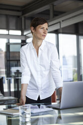 Geschäftsfrau stehend am Glastisch im Büro mit Laptop und Blick zur Seite - RBF06410