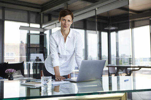 Porträt einer selbstbewussten Geschäftsfrau, die mit einem Laptop an einem Glastisch im Büro steht - RBF06413
