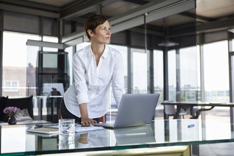 Geschäftsfrau stehend am Glastisch im Büro mit Laptop und Blick zur Seite, lizenzfreies Stockfoto