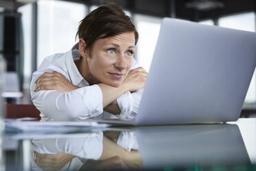 Businesswoman leaning on glass table in office looking at laptop - RBF06415