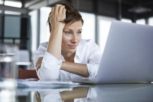 Frustrierte Geschäftsfrau sitzt am Glastisch im Büro und schaut auf den Laptop - RBF06416