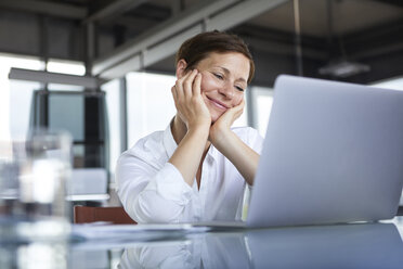 Lächelnde Geschäftsfrau, die an einem Glastisch im Büro sitzt und auf einen Laptop schaut - RBF06417