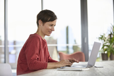 Lächelnde Frau sitzt am Tisch zu Hause und benutzt einen Laptop - RBF06419
