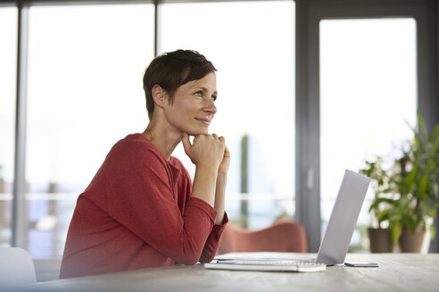 Lächelnde Frau sitzt am Tisch zu Hause und benutzt einen Laptop - RBF06420