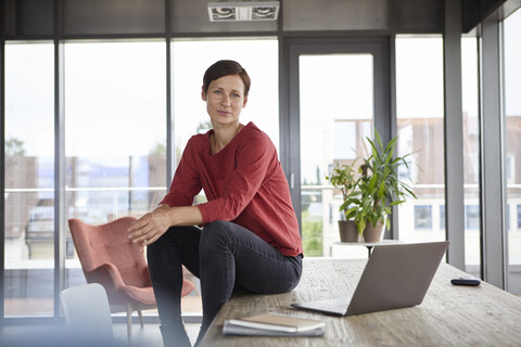 Portrait of confident woman sitting on table at home with laptop stock photo