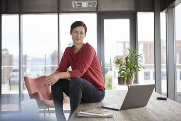 Portrait of confident woman sitting on table at home with laptop - RBF06422