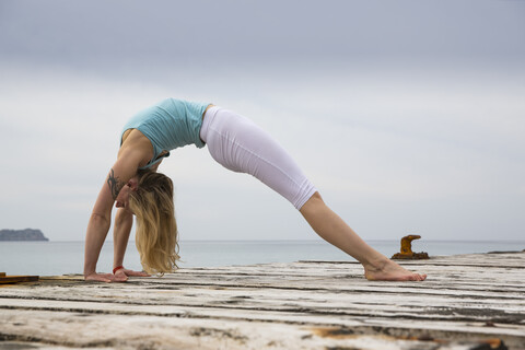 Mittlere erwachsene Frau, die sich nach hinten beugt und Yoga auf einer hölzernen Seebrücke praktiziert, lizenzfreies Stockfoto