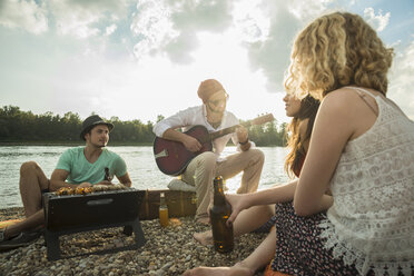Young man sitting by lake with friends playing guitar - CUF38136