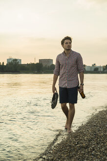 Young man walking by lake carrying shoes - CUF38142