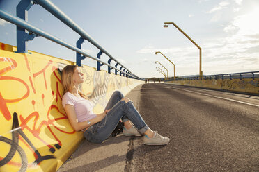 Junge Frau sitzt an eine Graffiti-Wand gelehnt auf einer Brücke im Sonnenlicht - CUF38570