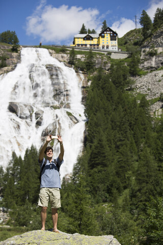 Junger Mann macht Smartphone-Selfie vor dem Toce-Wasserfall, Formazza, Verbania, Piemont, Italien, lizenzfreies Stockfoto