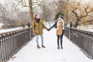 Junges verliebtes Paar steht Hand in Hand auf einer Fußgängerbrücke in einem Park an einem verschneiten Tag - WPEF00583