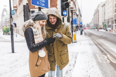 UK, London, young couple standing at roadside looking at cell phone on a snowy day - WPEF00590