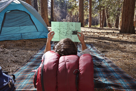 Rückansicht eines jungen männlichen Campers, der auf einer Decke liegt und auf eine Karte im Wald schaut, Los Angeles, Kalifornien, USA, lizenzfreies Stockfoto