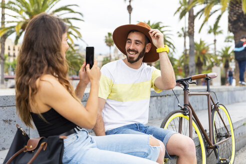 Spanien, Barcelona, glückliches Paar sitzt auf einer Bank und macht ein Smartphone-Foto - WPEF00623