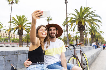 Spain, Barcelona, happy couple sitting on bench taking a selfie - WPEF00624