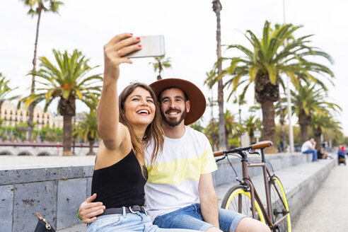 Spanien, Barcelona, glückliches Paar sitzt auf einer Bank und macht ein Selfie - WPEF00624