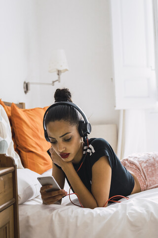 Schöne junge Frau, die zu Hause auf dem Bett liegt und Kopfhörer trägt und auf ihr Handy schaut, lizenzfreies Stockfoto