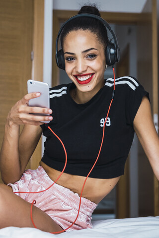Porträt einer lächelnden schönen jungen Frau, die mit Handy und Kopfhörern auf dem Bett sitzt, lizenzfreies Stockfoto