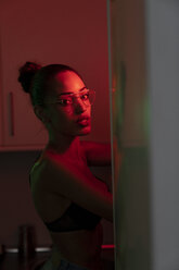 Porträt einer schönen jungen Frau mit BH und Brille in einem dunkel beleuchteten Raum - KKAF01140
