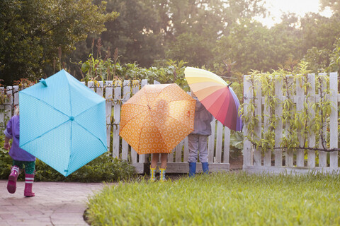 Junge und zwei Schwestern spielen im Garten und tragen Regenschirme, lizenzfreies Stockfoto
