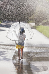 Rear view of barefoot girl carrying umbrella walking through street puddle - ISF16400