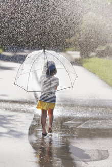 Rückansicht eines barfüßigen Mädchens, das einen Regenschirm trägt und durch eine Straßenpfütze läuft - ISF16400