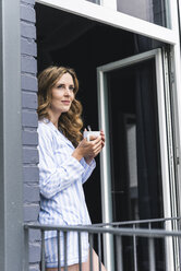 Lächelnde Frau im Pyjama zu Hause mit einer Tasse Kaffee und Blick aus der Balkontür - UUF14327