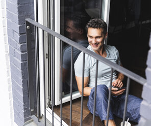 Lächelnder Mann im Pyjama zu Hause mit Handy und Blick aus der Balkontür - UUF14330