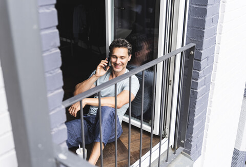Smiling man in pyjama at home on cell phone at balcony door stock photo