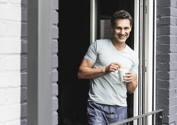 Portrait of smiling man in pyjama at home with cup of coffee at balcony door - UUF14332