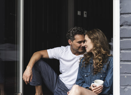 Affectionate couple in nightwear at home sitting at French window - UUF14339